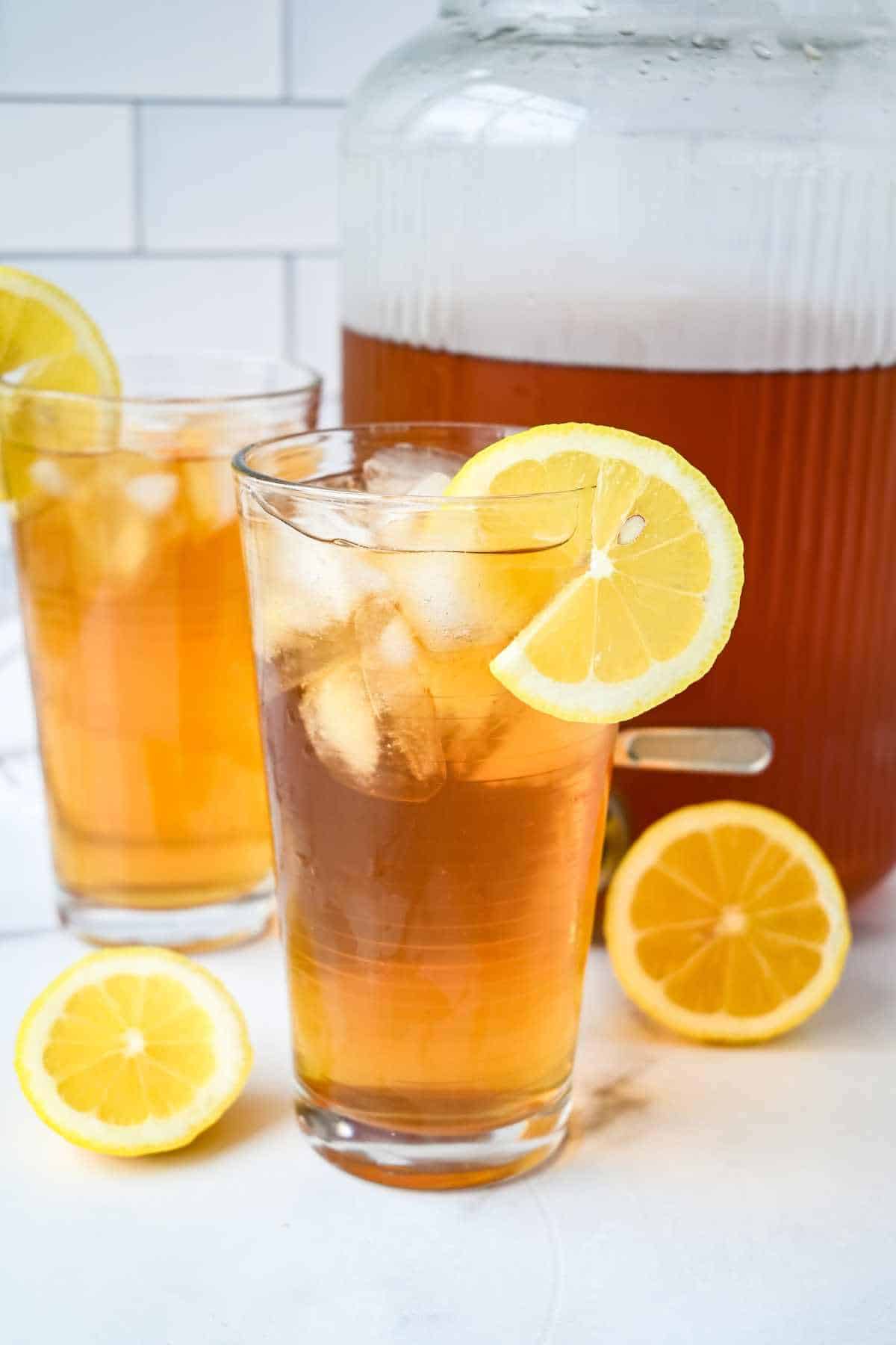 2 glasses of cold brew iced tea with lemons with a dispenser behind.