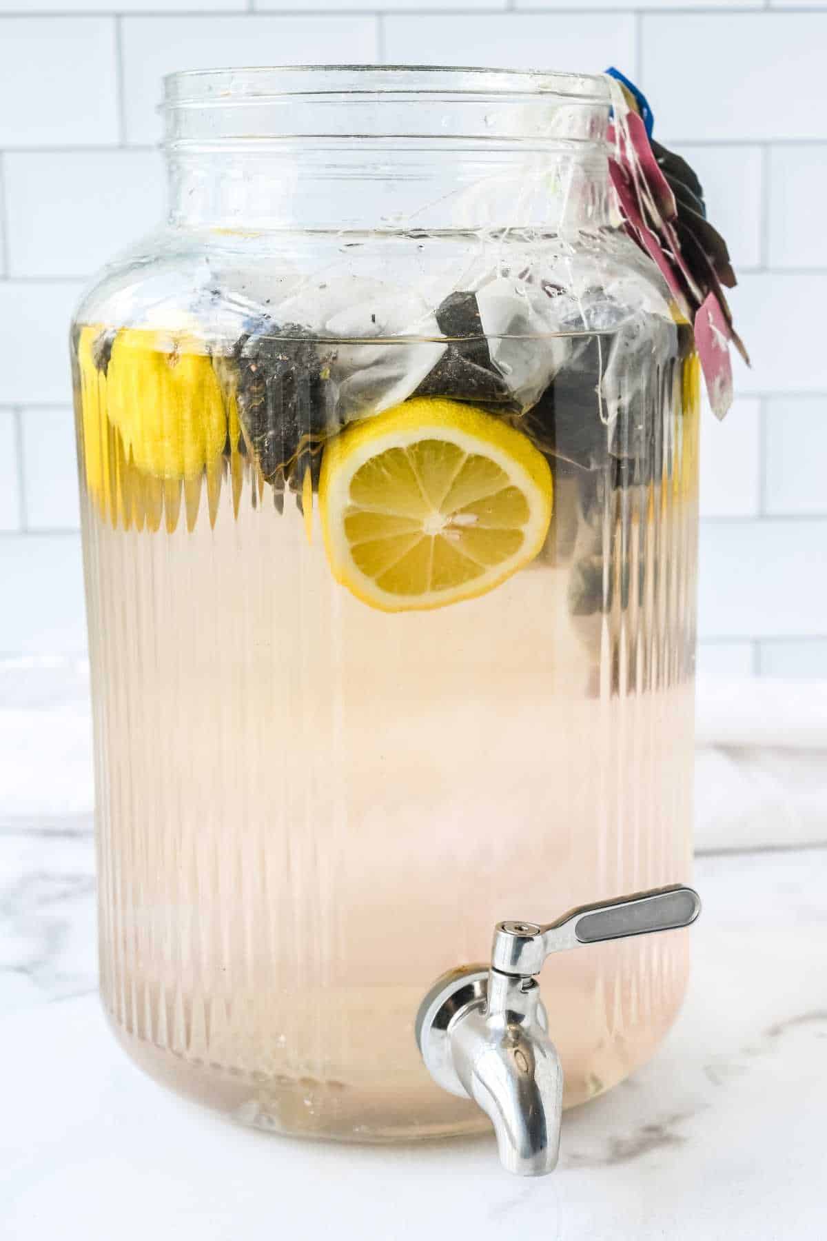 large glass beverage dispenser filled with water, lemons, and tea bags on a white background.