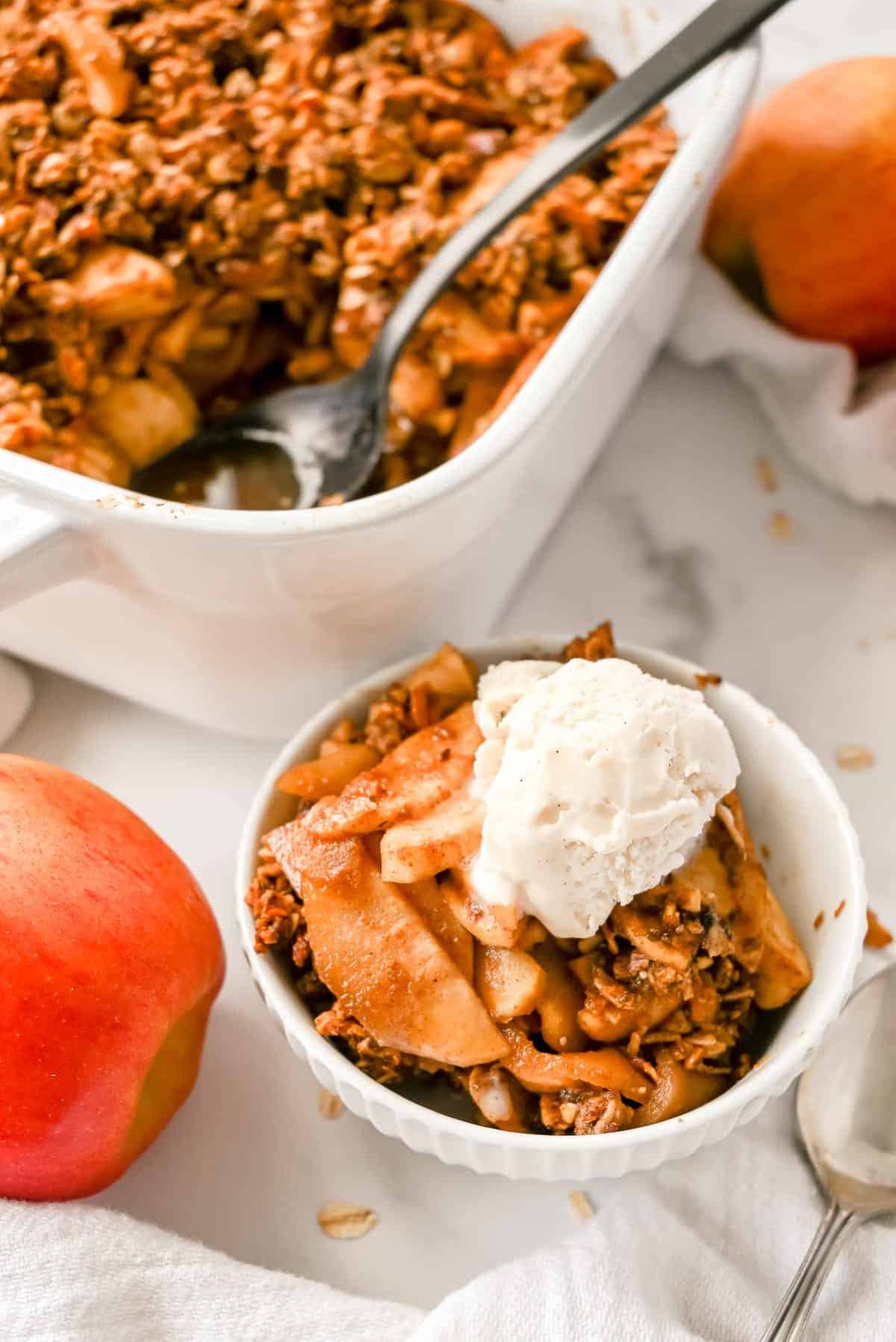 pan of healthy apple crisp with a spoon and a bowl topped with vanilla ice cream