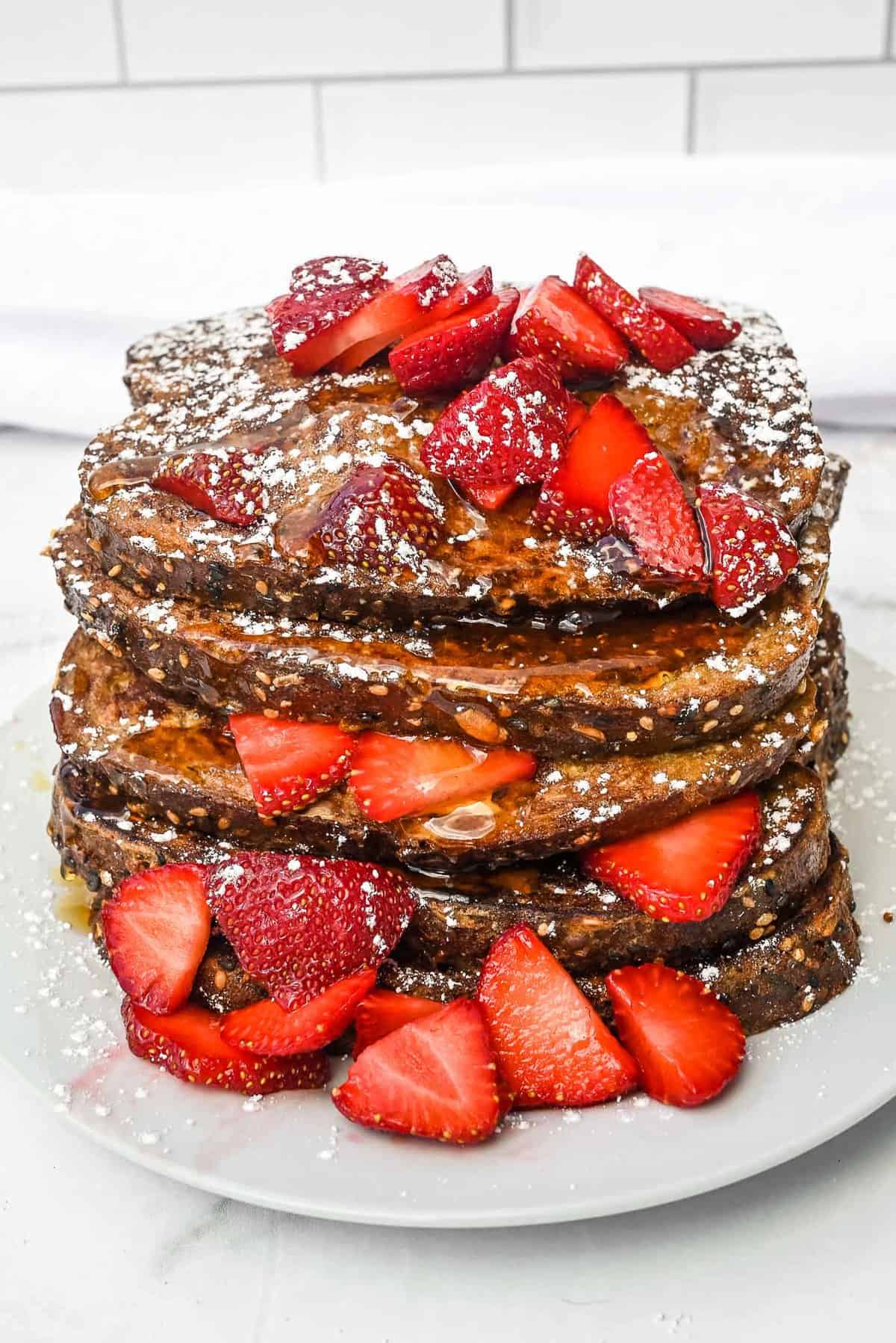 stack of dave's killer bread french toast topped with strawberries and powdered sugar on a white plate.