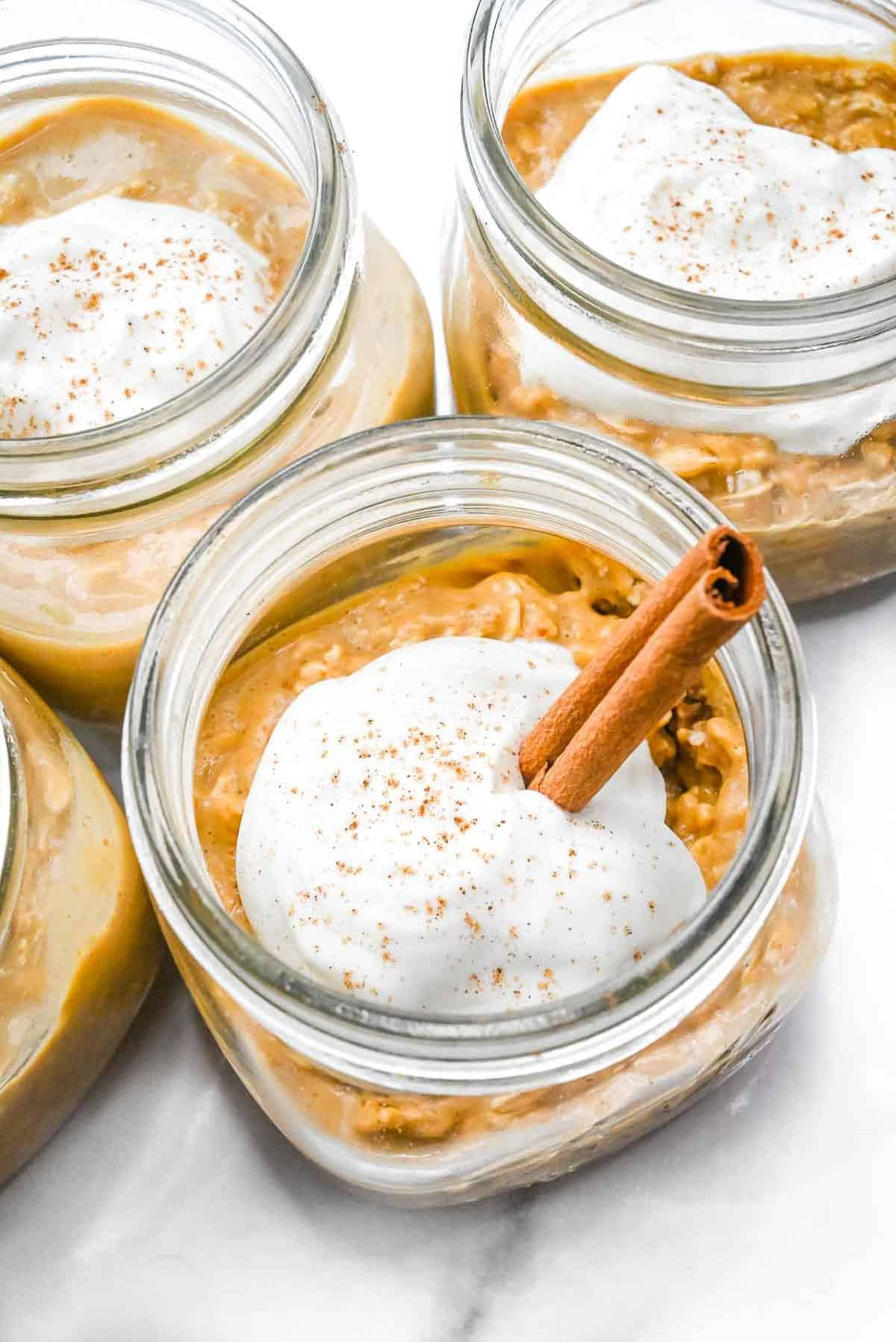 close up view of pumpkin spice overnight oats topped with yogurt and a cinnamon stick in a mason jar on a white background.