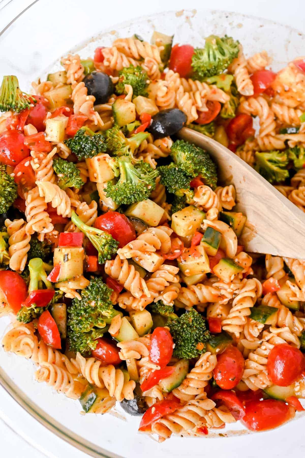 gluten-free pasta salad with vegetables and a wooden spoon in a glass bowl on a white background.