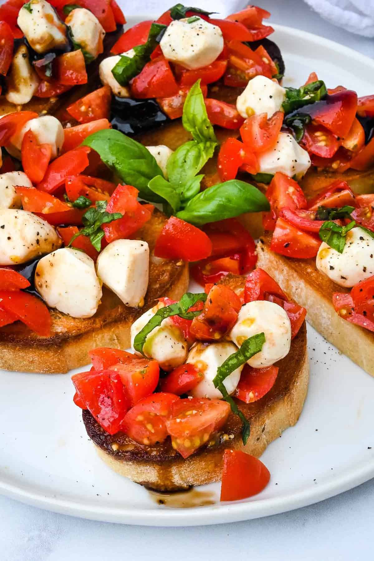toast topped with bruschetta, cheese and basil on a white plate.