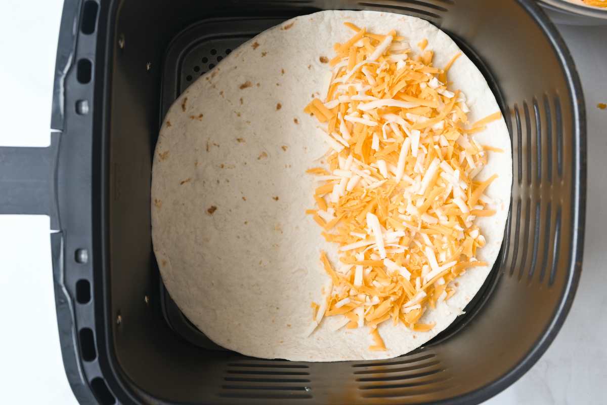 tortilla in air fryer basket with shredded cheese on one side.