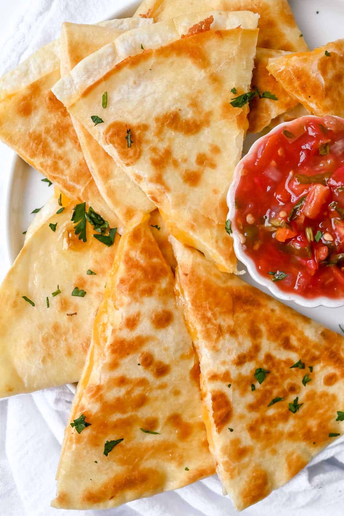 white plate of cheese quesadilla quarters topped with cilantro next to a bowl of salsa on a white background.