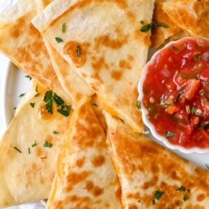 close up of white plate of cheese quesadilla quarters topped with cilantro next to a bowl of salsa on a white background.