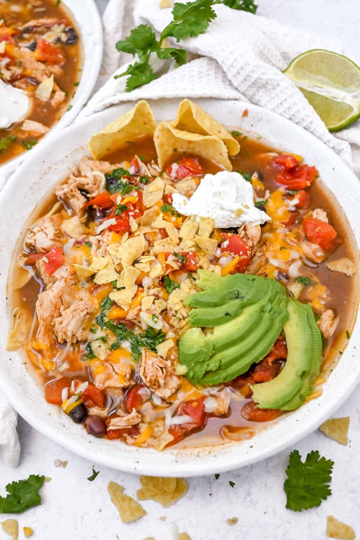 5-ingredient chicken tortilla soup in a white bowl topped with avocado and sour cream on a white background.