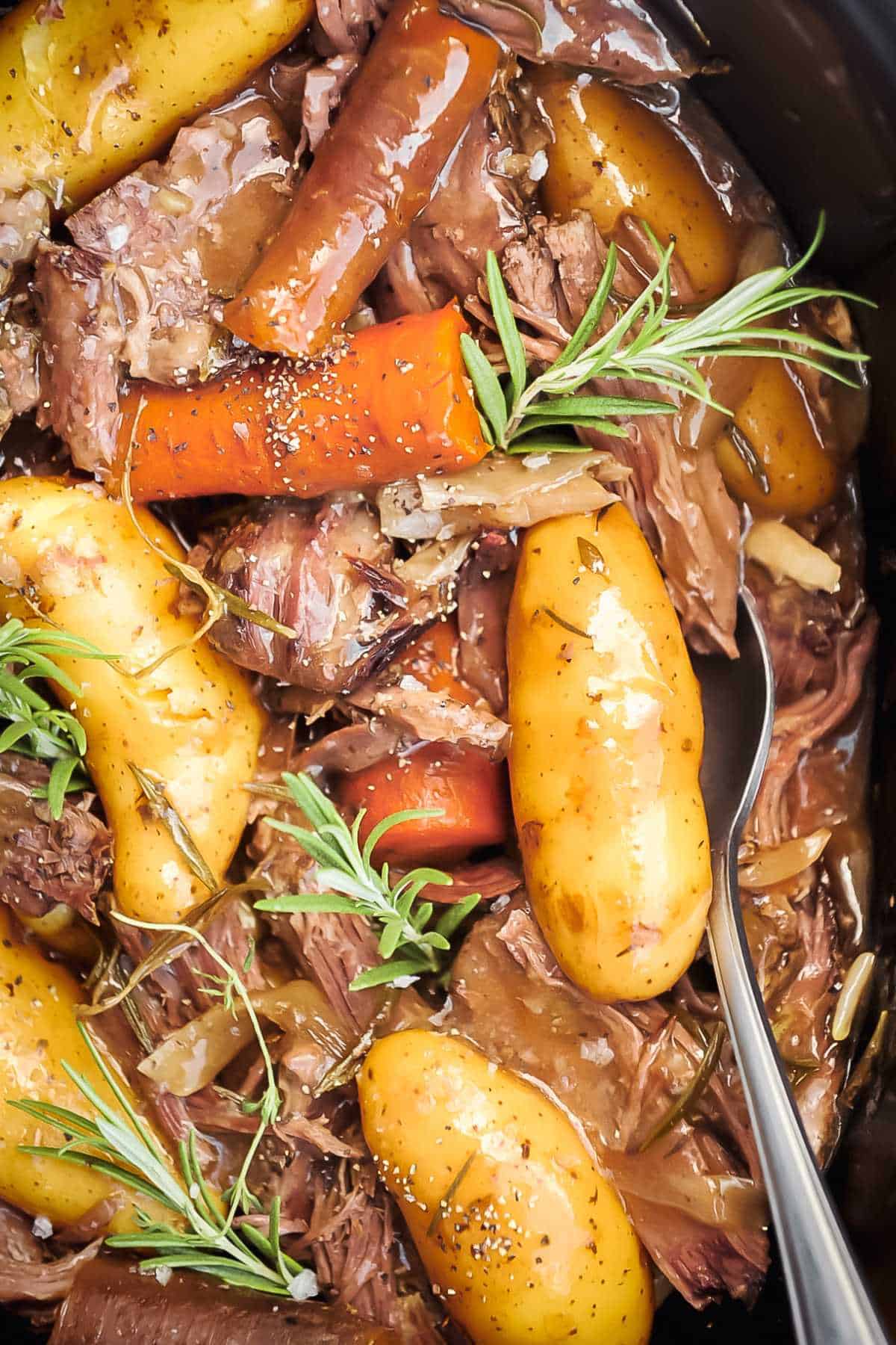 close up shot of crock pot roast with potatoes, carrots, and meat, topped with rosemary.