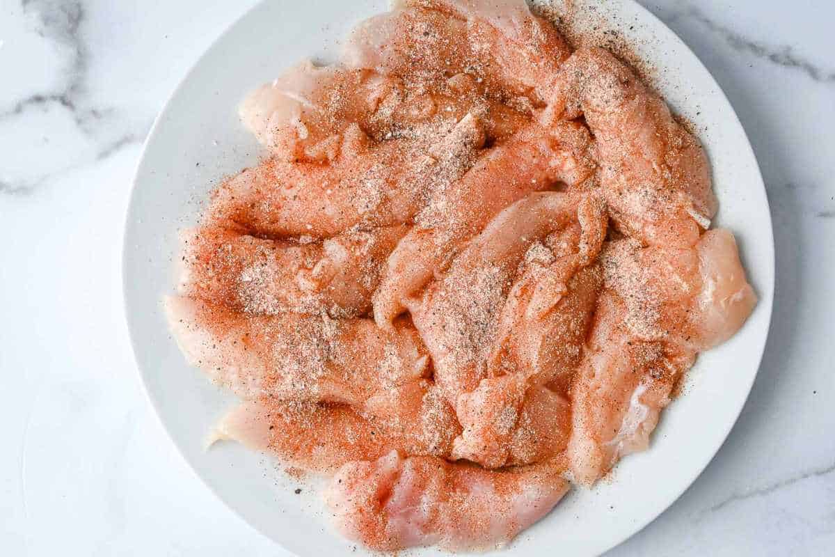 top down view of seasoned raw chicken tenders on a white plate on a white background.