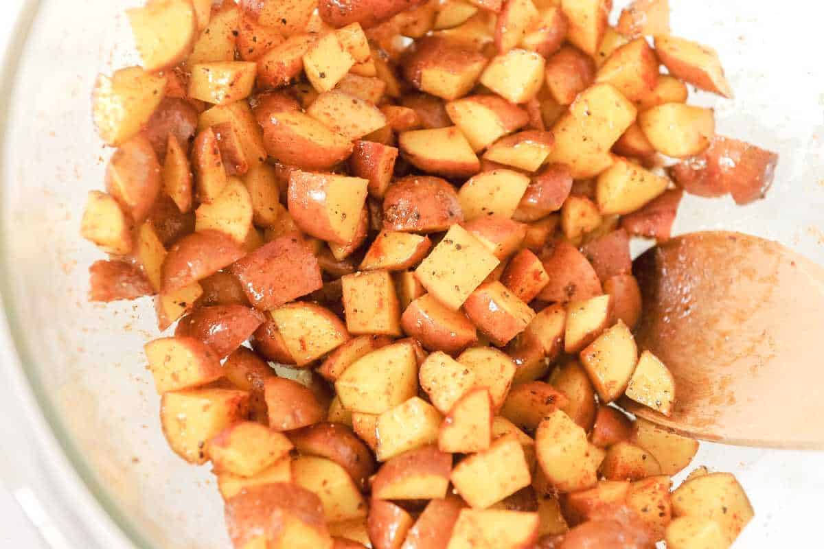 cubed breakfast potatoes with seasoning in a glass bowl with a wooden spoon.