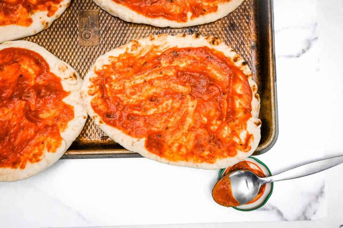 naan bread pizza topped with sauce on a sheet pan on a white marble background.