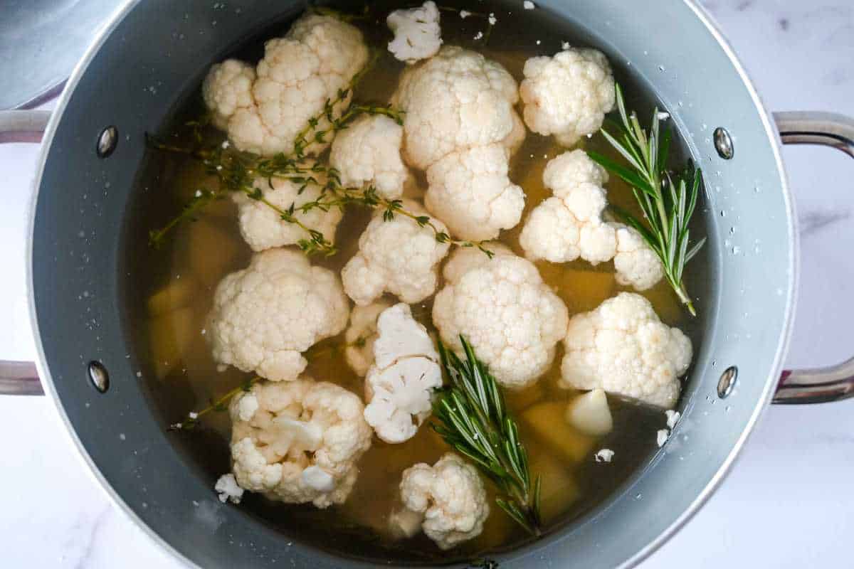 cauliflower, potatoes, and fresh herbs in water in a pot.