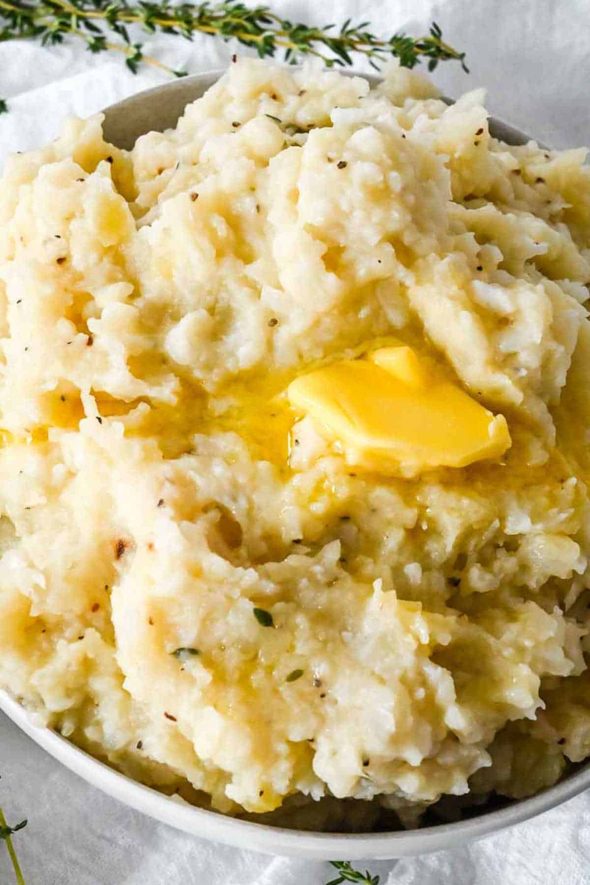 close up of mashed potatoes and cauliflower in a white bowl topped with butter.
