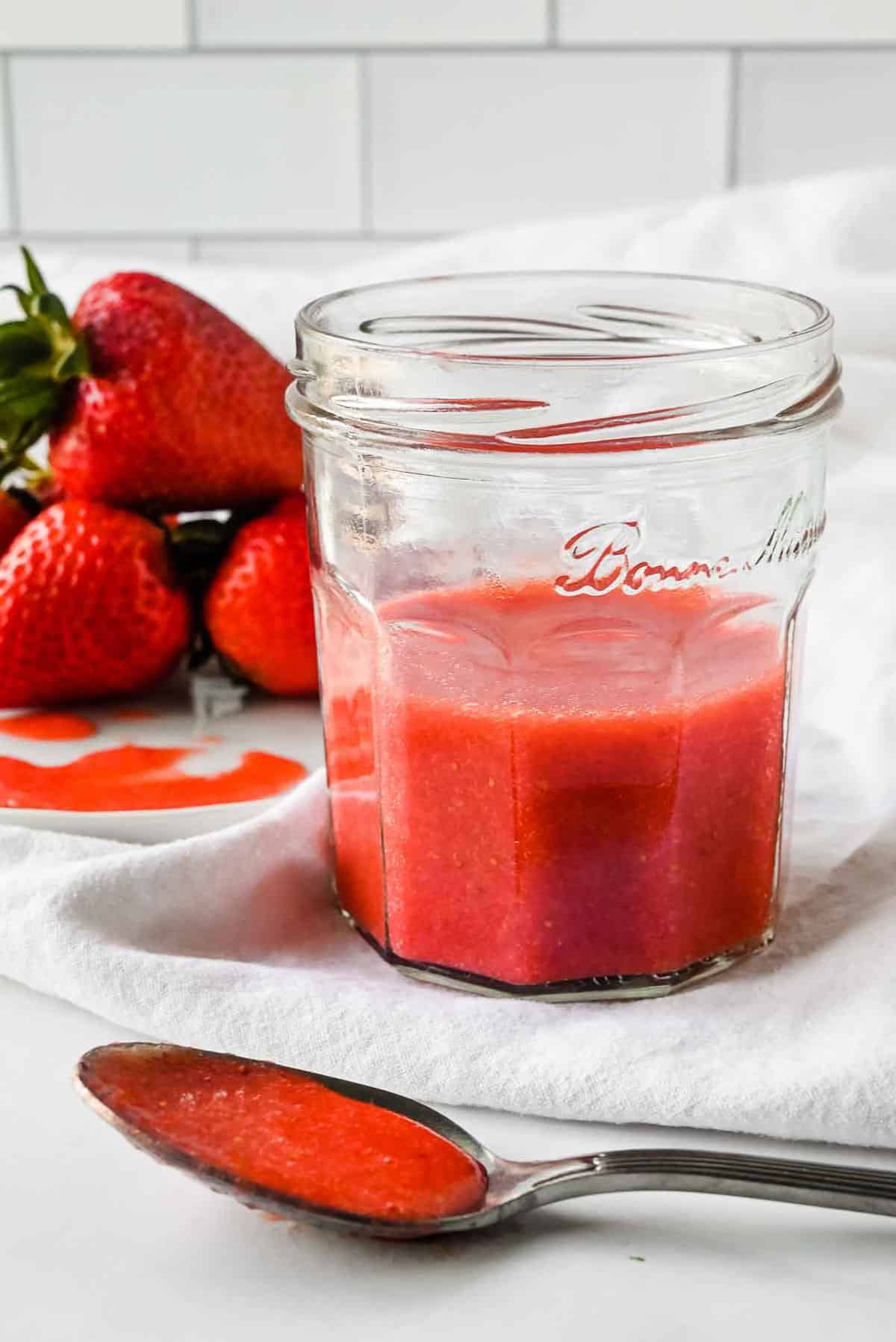jar of strawberry puree on a white napkin with strawberries in the background and a spoon with puree.
