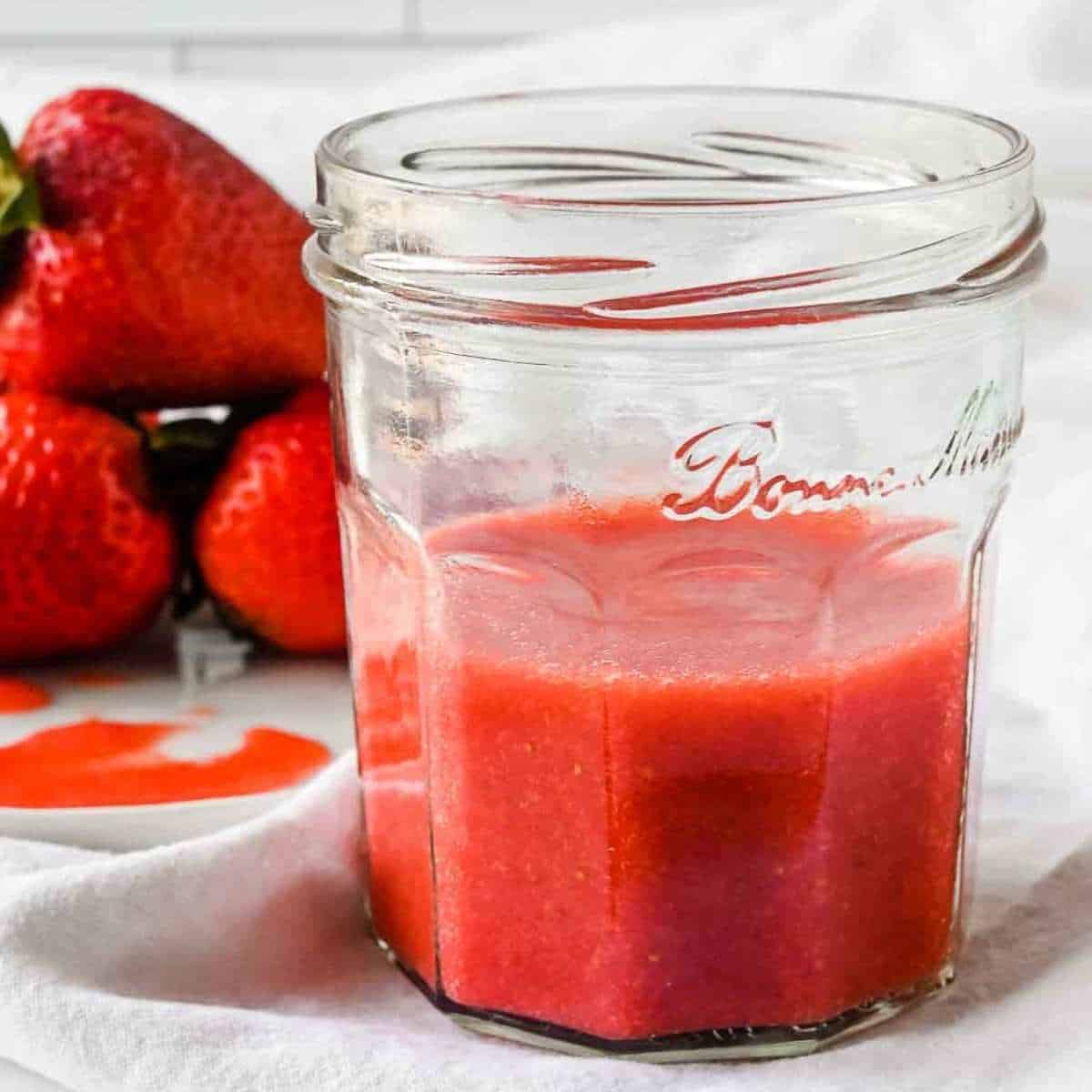 close up shot of strawberry puree in a jar with strawberries behind on a white background.
