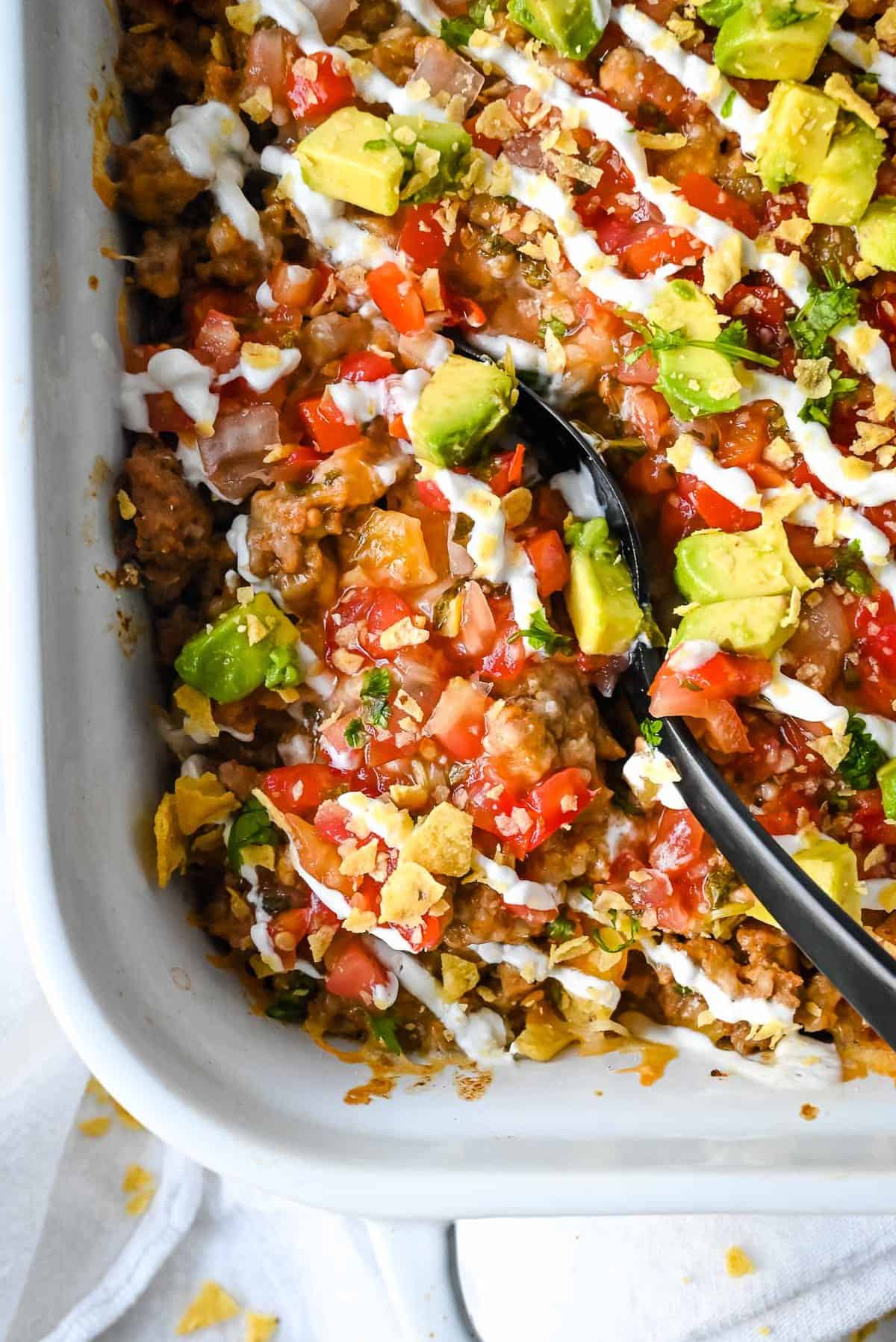 walking taco casserole bake topped with salsa, avocado and sour cream in a white casserole dish with a black spoon.