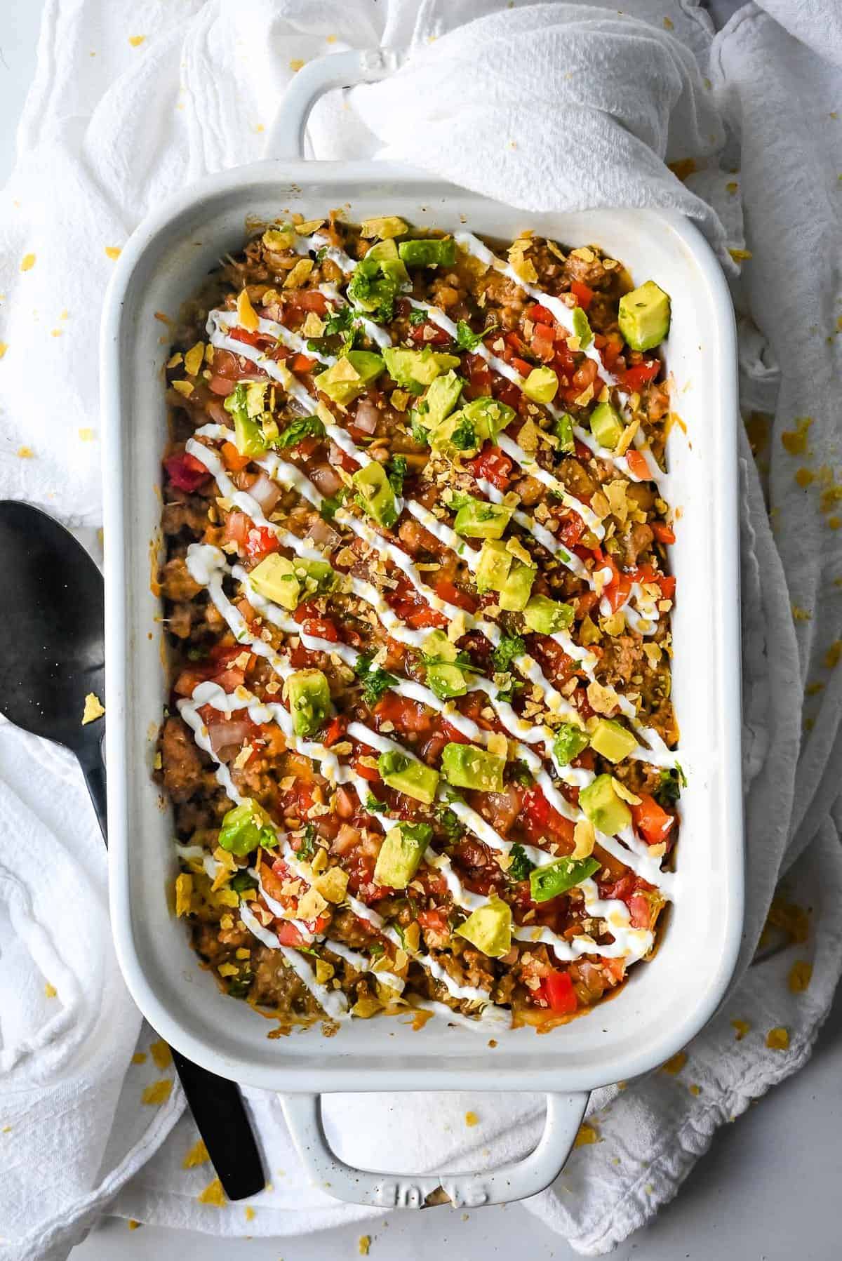 full shot of walking taco bake topped with avocado, cilantro, salsa and sour cream in a white casserole dish with a black spoon.