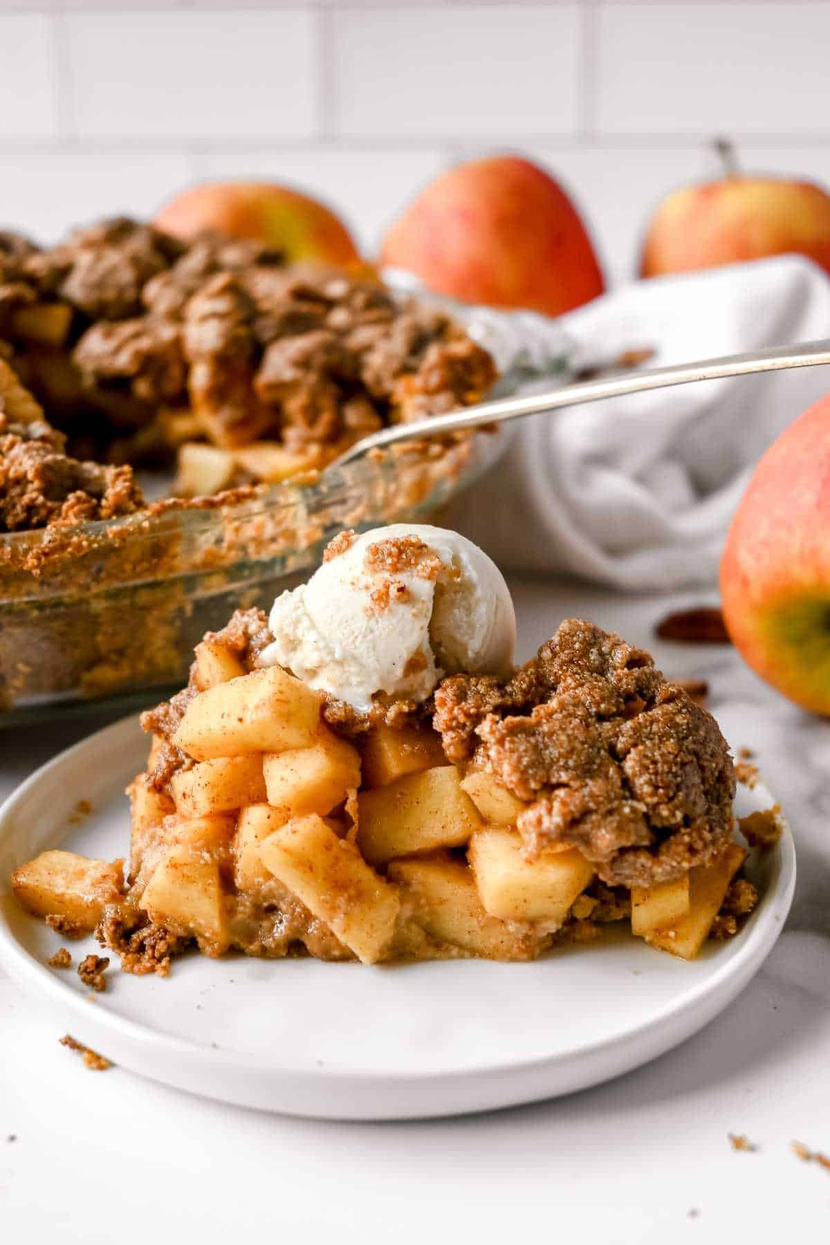 slice of healthy apple pie topped with ice cream on a white plate in front of the whole pie.