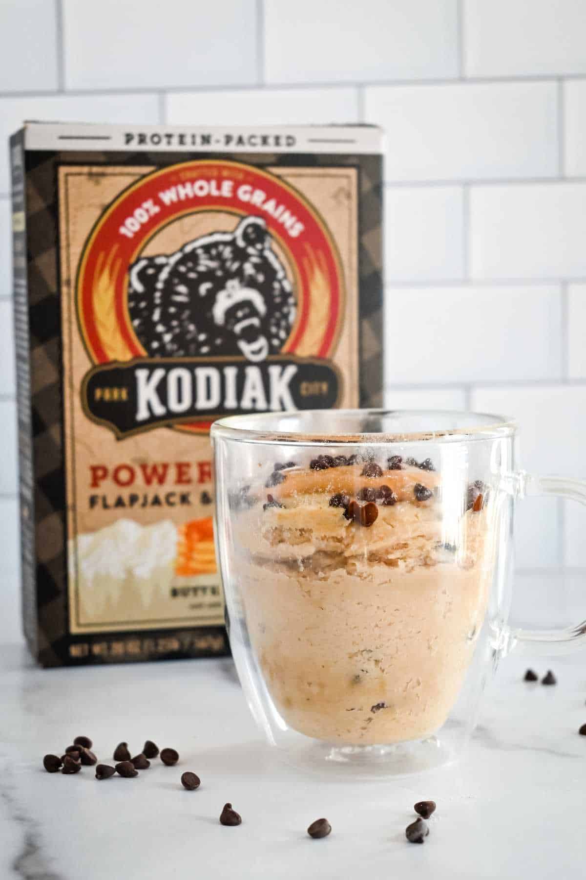 front view of kodiak cakes box behind pancake in a clear glass mug with chocolate chips and peanut butter.