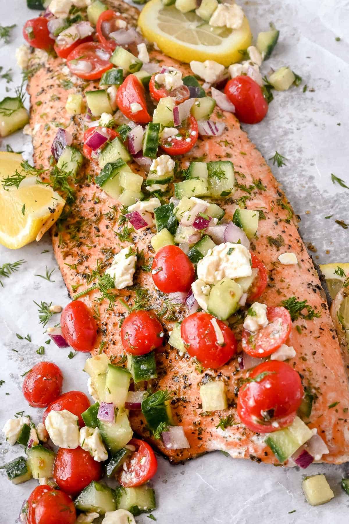 full shot of greek salmon fillet topped with cucumber, tomato, and feta salad with lemons on a white background.