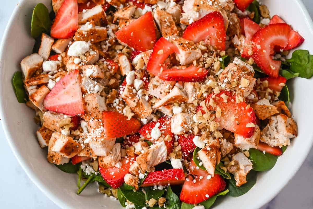 strawberries, walnuts, chicken, and goat cheese over spinach in a white bowl.