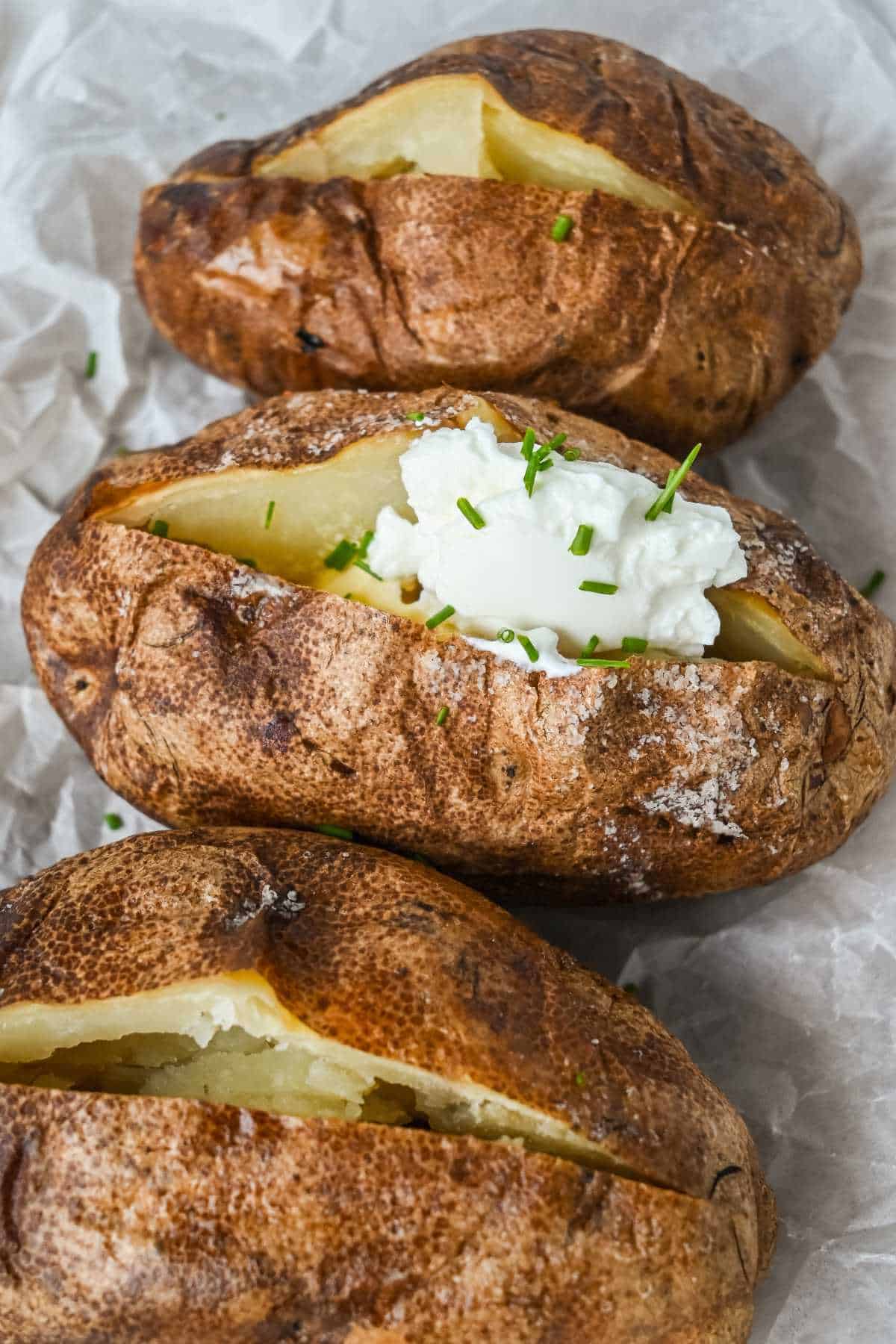 three baked potatoes split open with one topped with butter, sour cream, and chives.