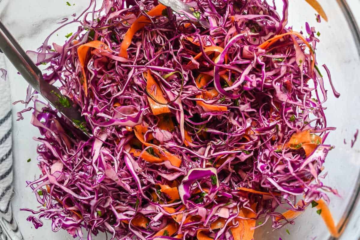 salad tongs mixing red cabbage slaw in a glass bowl.