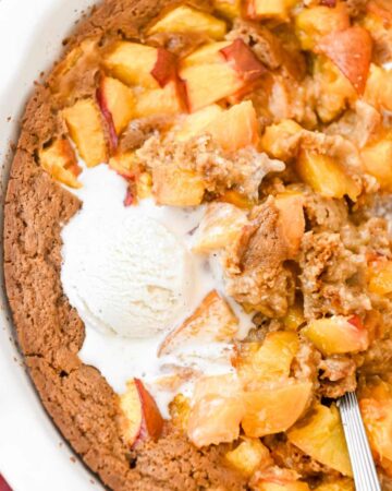 close up of nectarine cobbler in a white pie plate topped with vanilla ice cream with a spoon.