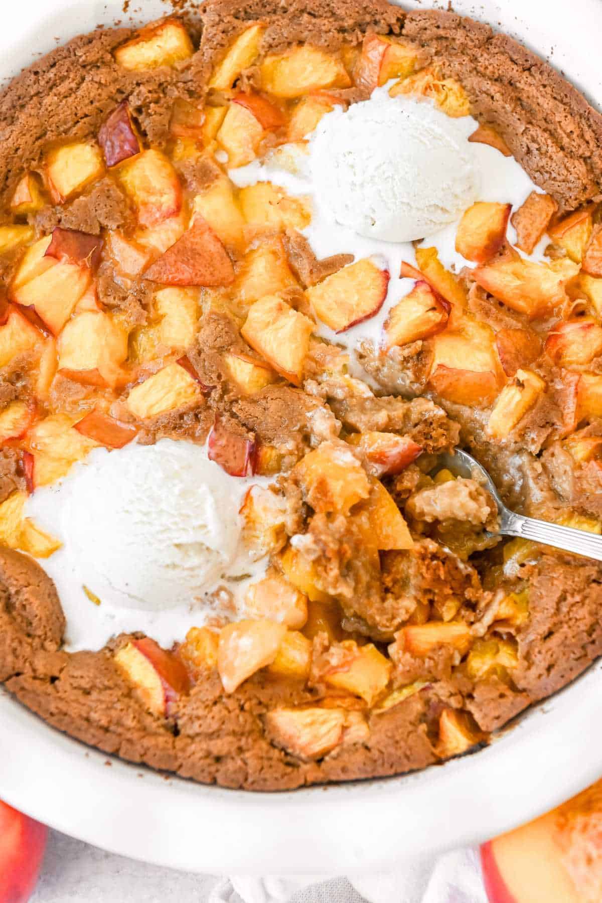 full shot of nectarine cobbler in a white pie plate with a spoon, topped with 2 scoops of vanilla ice cream.