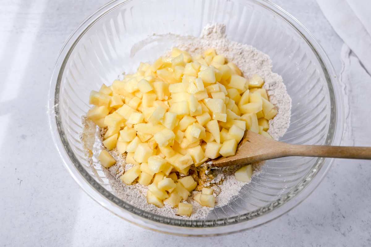 apples and dry ingredients in a glass bowl with a wooden spoon.