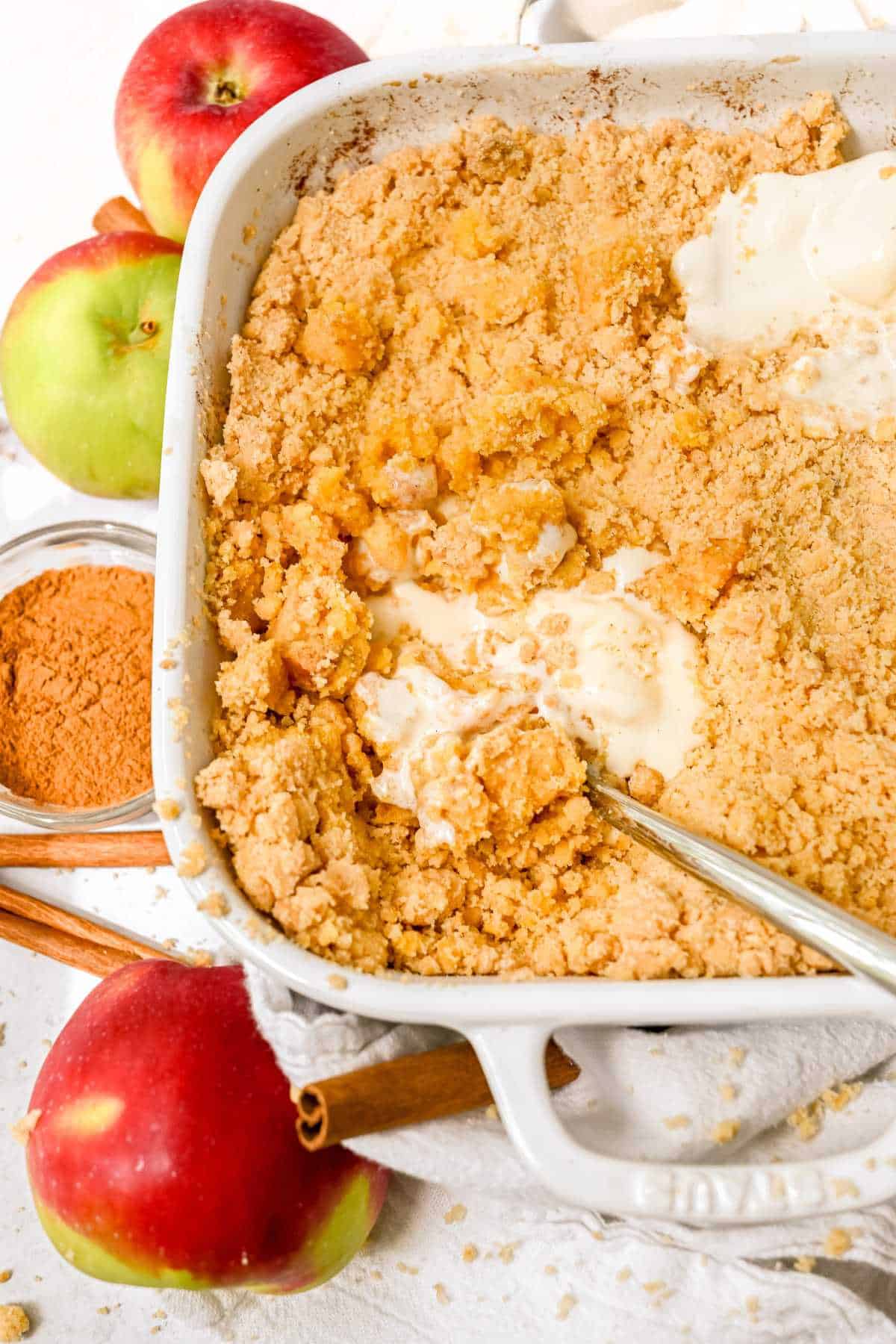 full view of cake mix apple cobbler in a white dish next to apples and cinnamon.