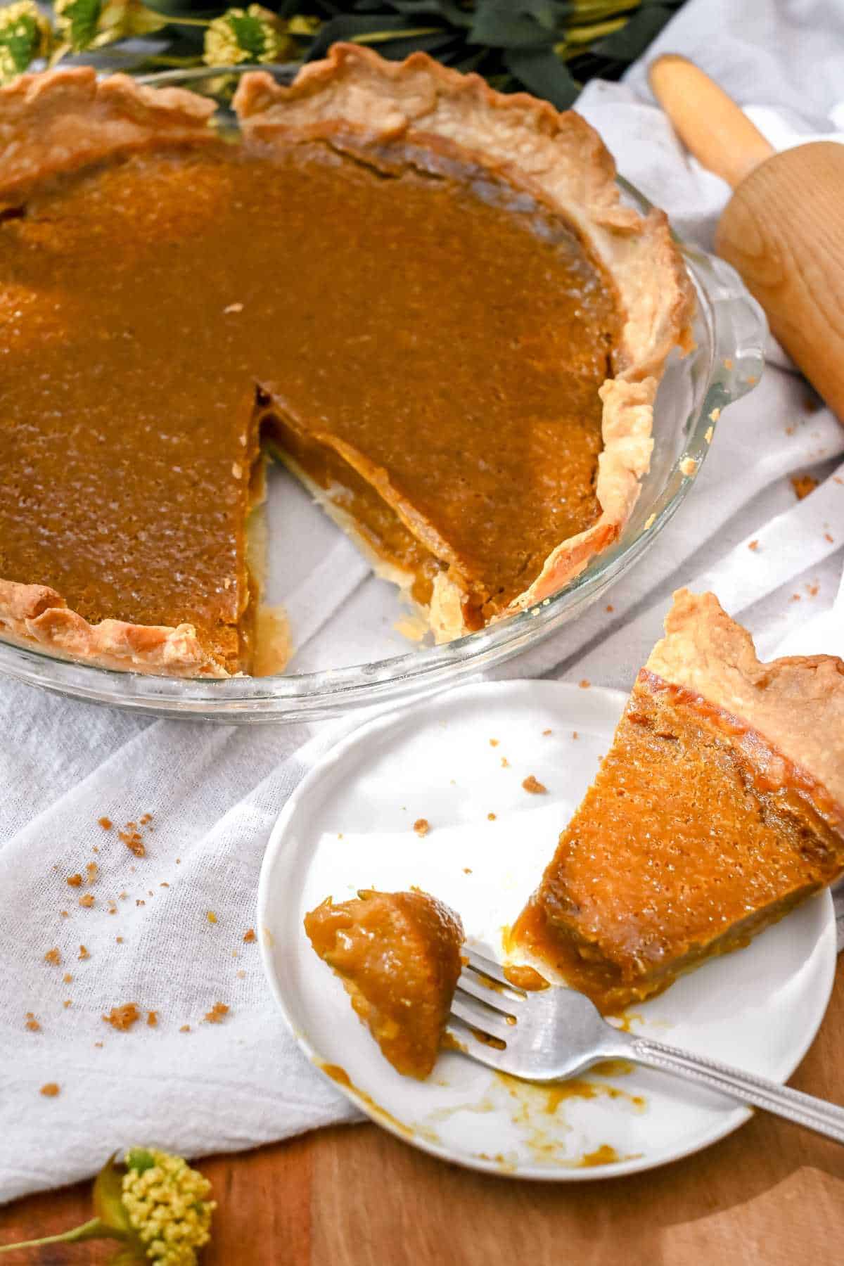 slice of pumpkin pie on a white plate with a fork next to a full pumpkin pie with one slice taken out.