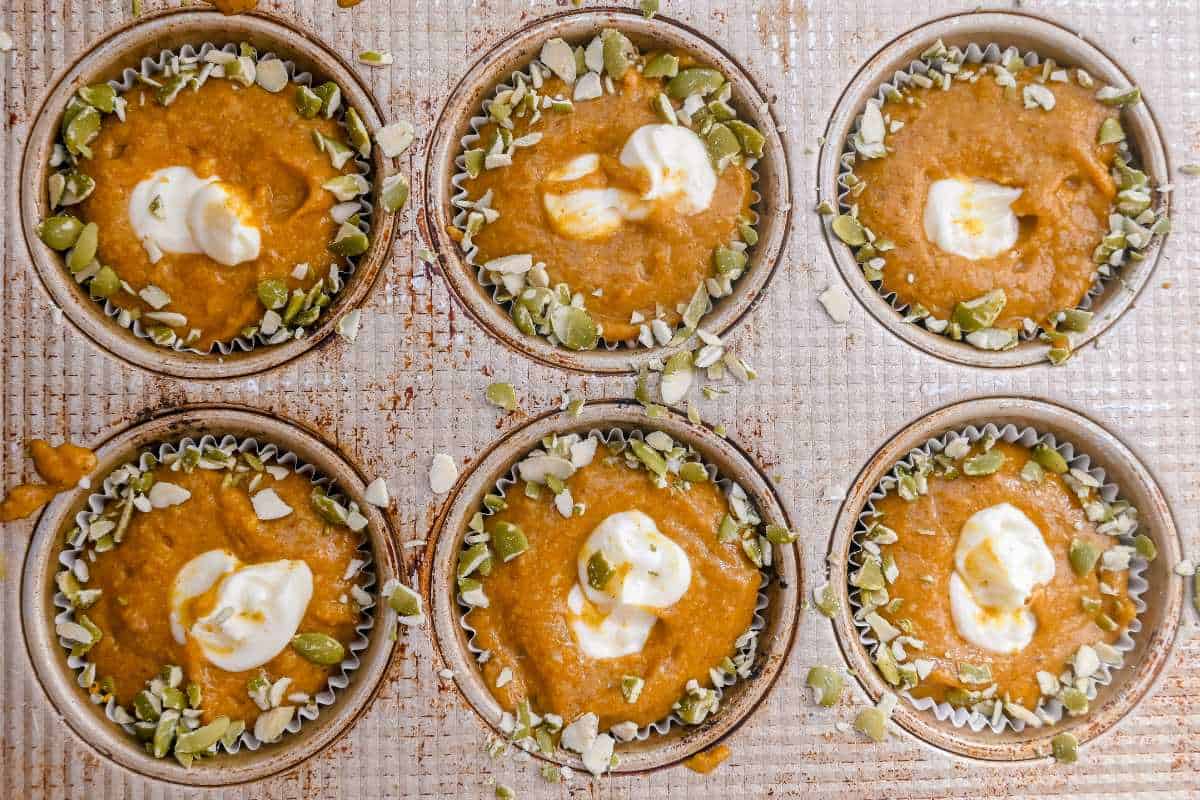 muffins in a pan unbaked and topped with cream cheese filling and pumpkin seeds.