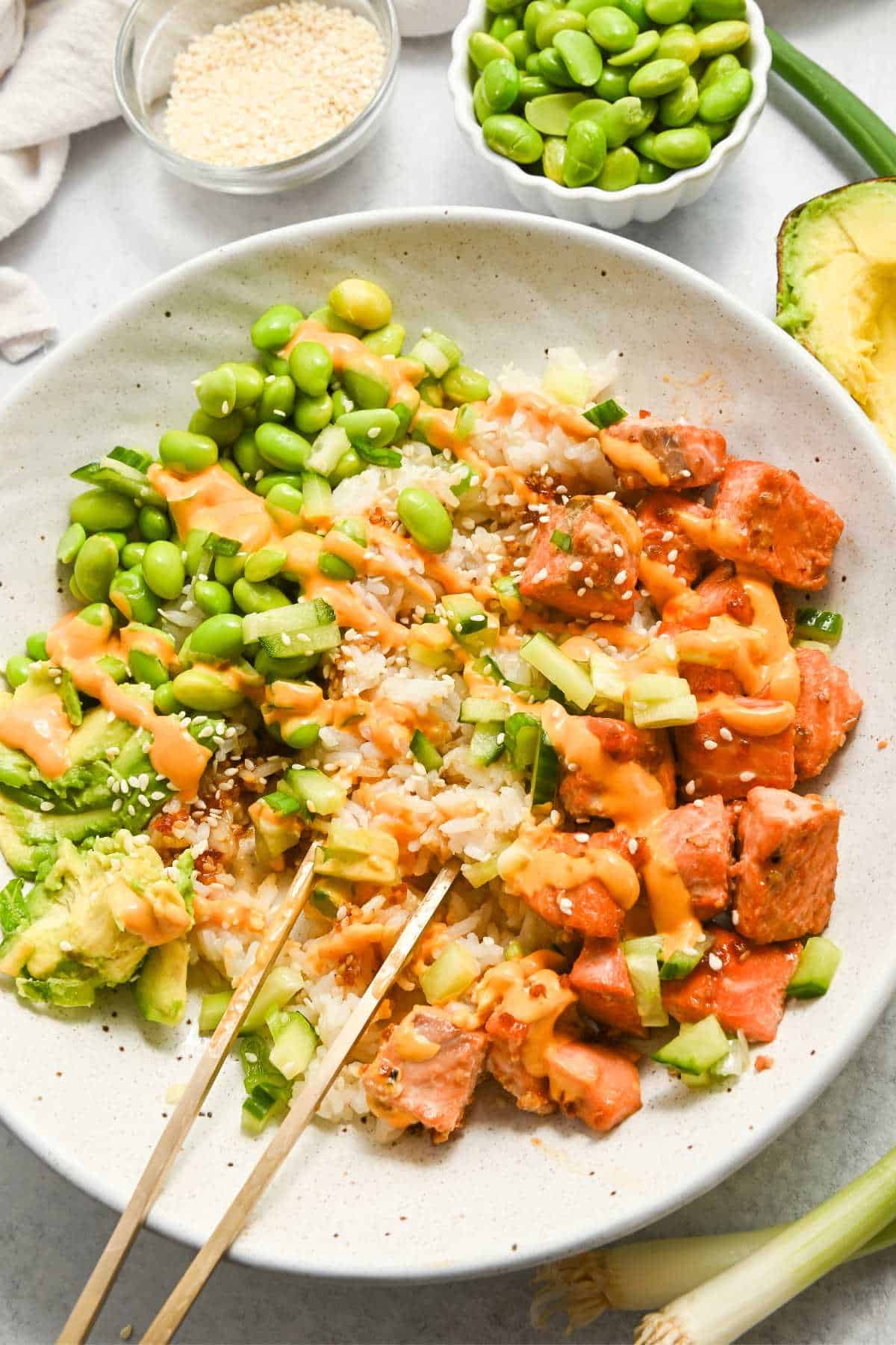 teriyaki salmon in a white bowl with rice, edamame, avocado, and with chopsticks on top.